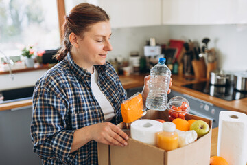 Young redhead woman with a delivery box full of fresh organic groceries in the kitchen. A donation box of different products on house background. Express food delivery, online shopping concept