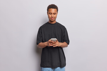 Horizontal shot of dark skinned man types text messages via smartphone uses contemporary technology dressed in casual black t shirt and jeans isolated over white background. Online communication