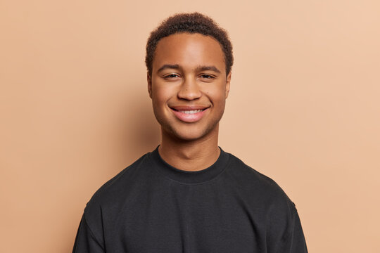 Cheerful young man with dark skin dressed casually in black tshirt smiles pleasantly looks directly at camera expresses genuine enjoyment isolated over brown background. People and emotions concept