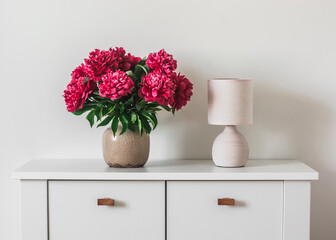 A bouquet of red peonies in a ceramic vase and a retro table lamp on a white chest of drawers. Minimalism style home interior