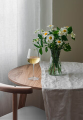 A glass of white wine on a round table with a bouquet of daisies and jasmine in a cozy living room