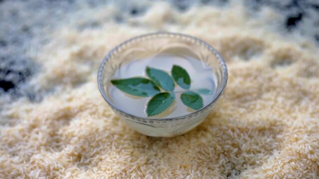 Oryza sativa and Oryza glaberrima or raw lachkari kolam rice grains on a black surface along with a glass bowl containing rice water in it with some rose leaves on top, used in beauty products and fac