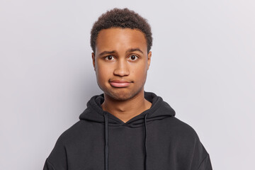 Obraz premium Portrait of handsome displeased man purses lips has worried expression focused at camera hears something unpleasant wears black sweatshirt isolated on white background. Negative human face expressions