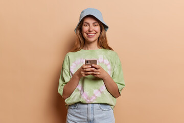 Indoor shot of good looking woman uses cell phone messages being happy to text with friends wears panama t shirt and jeans isolated over brown background. People and modern technologies concept