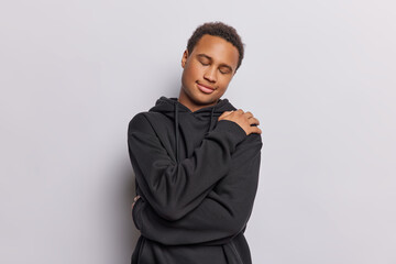 Obraz premium Horizontal shot of dark skinned man wraps his arms around body keeps eyes closed has gentle smile on face feels comfortable dressed in casual black sweatshirt isolated over white background.
