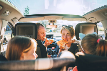 Young couple with daughters eating just cooked Italian pizza sitting in modern car with transparent...