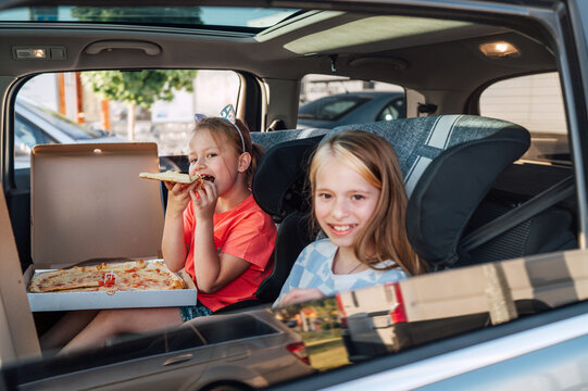 Two positive smiling sisters are happy to eat just cooked Italian pizza sitting in child car seats on the car back seat. Happy childhood, fast food eating, or auto journey lunch break concept image.