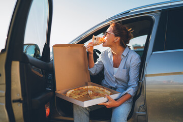 Midle-aged woman eating just cooked italian pizza sitting on driver car seat during meal break and...