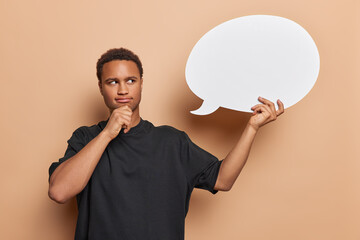 Thoughtful young man holds chin and looks pensively at blank speech bubble considers something what...