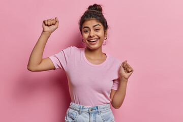 Energetic carefree Indian girl has fun shakes arms enjoys favorite music smiles broadly wears casual t shirt and jeans poses against pink wall spends great time at friends party being full of energy - 609665903
