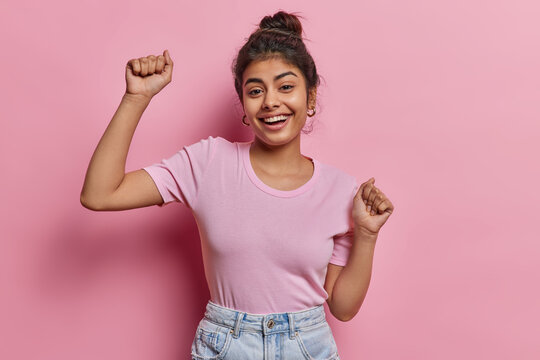 Energetic carefree Indian girl has fun shakes arms enjoys favorite music smiles broadly wears casual t shirt and jeans poses against pink wall spends great time at friends party being full of energy