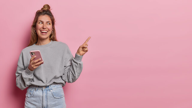 Horizontal shot of good looking positive young woman with hair bun dressed in casual ;clothes uses mobile phone for chatting online points index finger aside on blank space to place your advertisement
