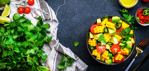Vegan salad with sweet corn, avocado, jalapeno pepper, red tomatoes, fresh cilantro and olive oil and lime dressing. Black table background, top view
