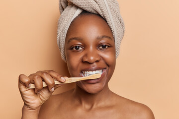 Healthy morning routine. Dark skinned woman showcases commitment to oral hygiene by diligently brushing teeth using toothbrush and toothpaste stands shirtless isolated over brown background.