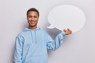 Pleased delighted young African man holds empty speech bubble with mock up space for text dressed in casual blue hoodie suggests to place your advertisement here stands against white background.