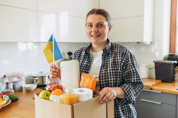 Female Volunteer holding box with groceries and flag of Ukraine on the house kitchen background. Donation box with food in female hands. Concept of humanitarian, donations or helping