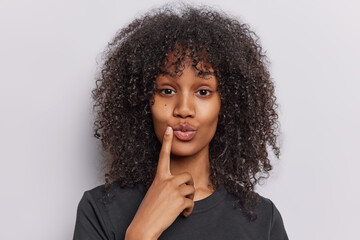 Fototapeta na wymiar Portrait of lovely millennial girl with curly bushy hair keeps finger near folded lips has friendly face expression dressed in casual black t shirt isolated over white background. Studio shot