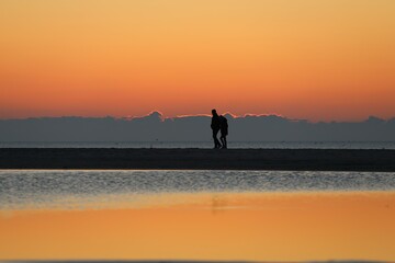 Fototapeta premium Liebespaar geht am Strand entlang im Abendrot hinter Spiegelungen im Wasser