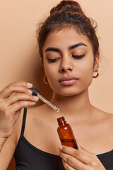 Serious dark haired young woman holds bottle with nourishing serum to face looks attentively...