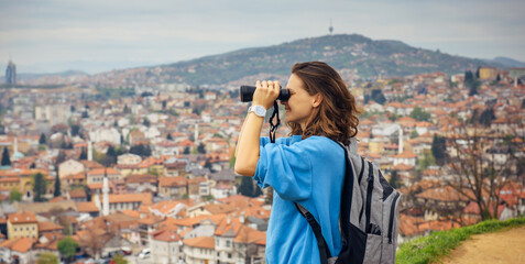 A beautiful girl traveler looks through binoculars at Sarajevo, the capital of Bosnia and Herzegovina. View of the Balkan European city with orange roofs in the mountains from a view point