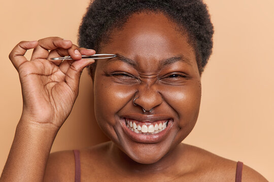 Close up shot of cheerful young dark skinned woman plucks eyebrows with tweezers enhances natural beauty engaged in self care tenderly sculpting face clenches teeth poses indoor against brown wall