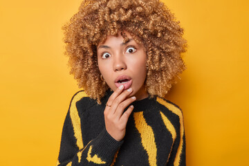 Photo of impressed stunned curly haired young European woman keeps hand on lips has mouth widely...