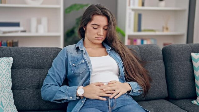 Young beautiful hispanic woman suffering for stomach ache sitting on sofa at home