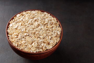Oat flakes in a clay bowl on a dark background. Organic rolled oats for healthy nutrition. Copy space. Top view