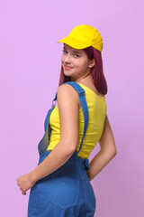 A girl in a blue denim overalls and a yellow T-shirt and a baseball cap on a pink studio background