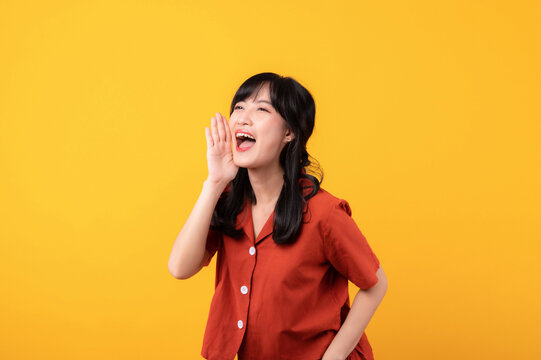 Portrait young beautiful asian woman happy smile dressed in orange clothes shout to free space isolated on yellow studio background. Presenting product Beautiful girl act like telling to customer
