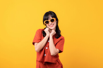 Portrait young beautiful asian woman cute and shy dressed in orange clothes and sunglasses touching cheeks showing shy emotional feeling isolated on yellow studio background.