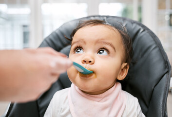 Adorable, sweet and cute baby eating puree for lunch, dinner or snack in her high chair at home. Child development, food and girl infant kid enjoying meal with spoon for growth and wellness in house.