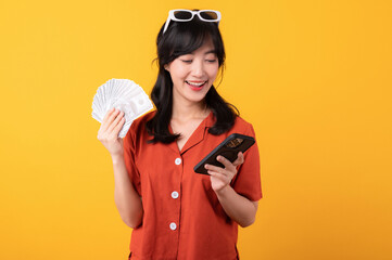 Portrait young beautiful asian woman happy smile dressed in orange clothes showing cash money and smartphone isolated on yellow background. Pay and purchase shopping online payment concept.