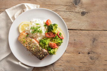 Crispy fried salmon filet with vegetables from broccoli and tomato, rice, parsley and lemon zest on a rustic wooden table, copy space, high angle view from above