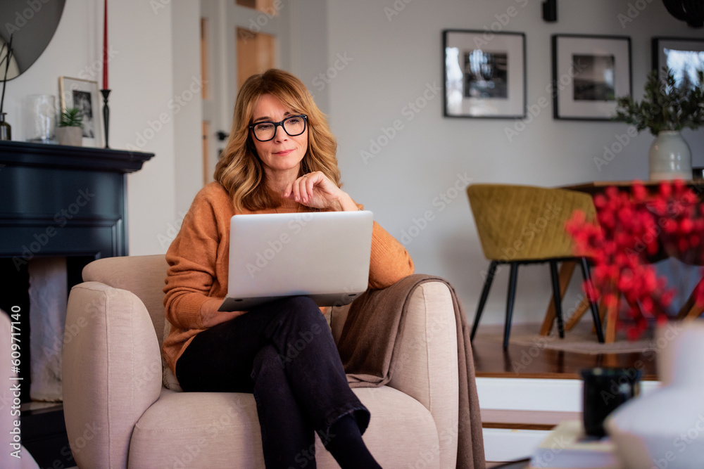 Sticker Mid aged woman sitting in a modern home and using laptop for work