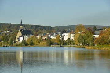 Fototapeta na wymiar Le lac des Doyards et le village de Vielsalm avec l'église paroissiale Saint Gengoux se reflétant dans ses eaux sous un ciel clair d'automne 