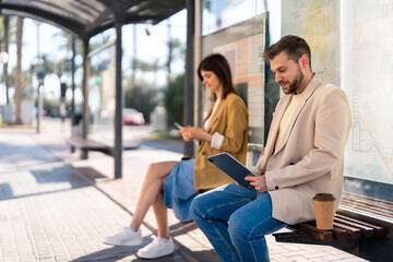 Confident business man with takeaway coffee sitting at bus station with a female, waiting for bus, using digital tablet device to check bus schedules, connections and maps, buying online bus ticket.