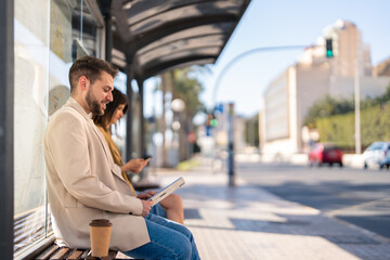 Side view of handsome young business man with beard in beige suit sitting at bus station, working on digital tablet or reading news, communicating online while waiting for bus in the city. Copy space.