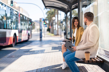 Two cheerful friends young woman and man sitting at bus station while city bus is passing by on the street, having a conversation, laughing, holding mobile phone and digital tablet. Copy space photo. - obrazy, fototapety, plakaty