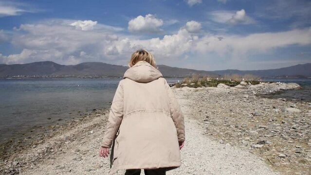 An adult blonde woman walking along the seashore runs and scares the sitting seagulls flying up into the sky. Cheerful happy woman on an autumn day off on the shore of a mountain lake