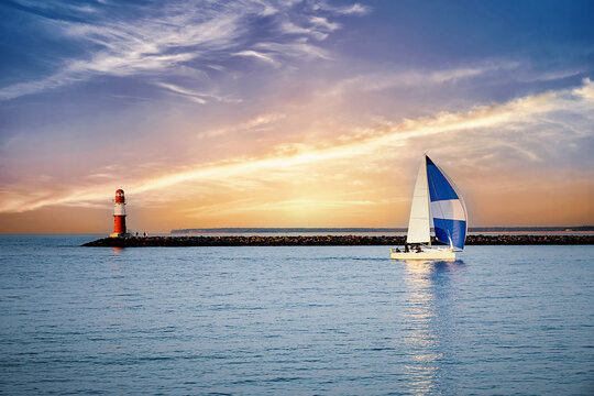 Sailing ship off the coast of the Baltic Sea with a red lighthouse and spectacular sky features of sunshine and clouds.