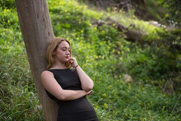 Young woman, blonde and beautiful, with a black dress, leaning on the trunk of a tree, in the middle of nature, alone, pensive and calm. Concept nature, thoughts, solitude, calm, trees.