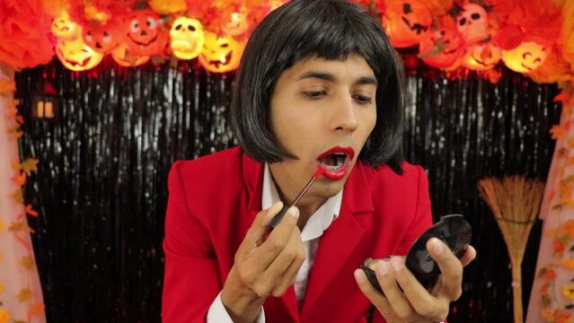 Man disguised as a woman putting on makeup at halloween party. Transvestite, transexual. Red jacket, microphone, black wig. He paints his lips.
