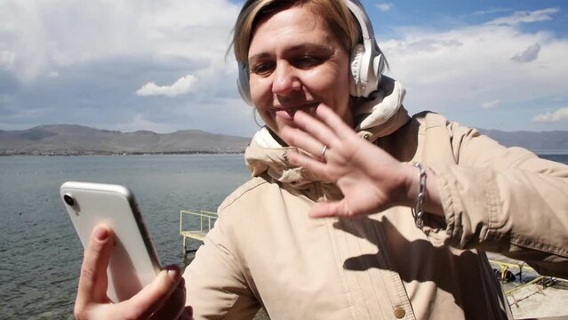 Adult young woman talking on a video call using a mobile phone and headphones standing on the seashore dressed in a jacket on a sunny autumn but windy day Travel and vacation