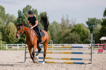 Female rider on chestnut horse jumping over a hurdle at the equestrian center on a sunny summer day. Equitation sport concept.