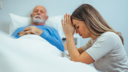 Obraz premium A woman praying at the bedside of a sick man in the hospital. The man is asleep in the background. He is wearing a blue pajama top. Her hands are clasped in front of her head