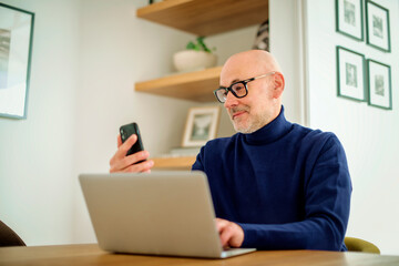 Middle aged businessman using smartphone and laptop while working from home