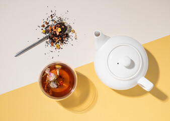 Creative flat lay of a glass cup of herbal tea with rose flowers on a yellow and white background with teapot and hard shadow. Food and drink concept.