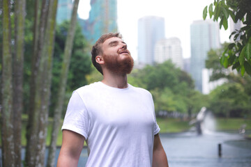 Positive young man smiling during the rain in the park. Cheerful male enjoying the rain outdoors. Guy looking up and catching the rain drop with hands. Breath deep and relax