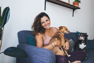 Portrait of happy Caucasian woman in casual clothing hug favourite dogs feeling love emotions during daily routine in home living room, cheerful hipster girl 20s take care of cute pets in collar